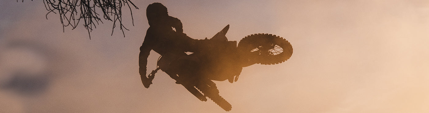 Silhouette einer Person, die auf einem Motocross-Motorrad vor einem nebligen, goldenen Himmel einen Stunt in der Luft ausführt. Vereinzelte Äste rahmen die Szene ein.