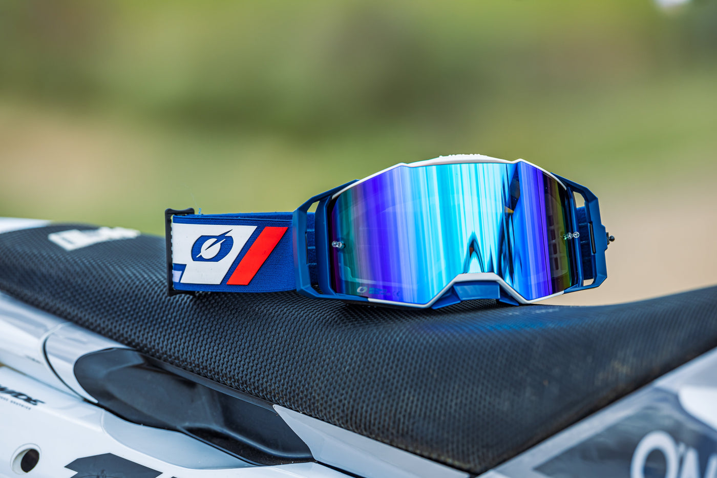 Eine blau-weiße Motocross-Brille mit bunten reflektierenden Gläsern liegt auf einem Motocross-Sitz. Der unscharfe Hintergrund lässt auf eine Outdoor-Umgebung schließen.