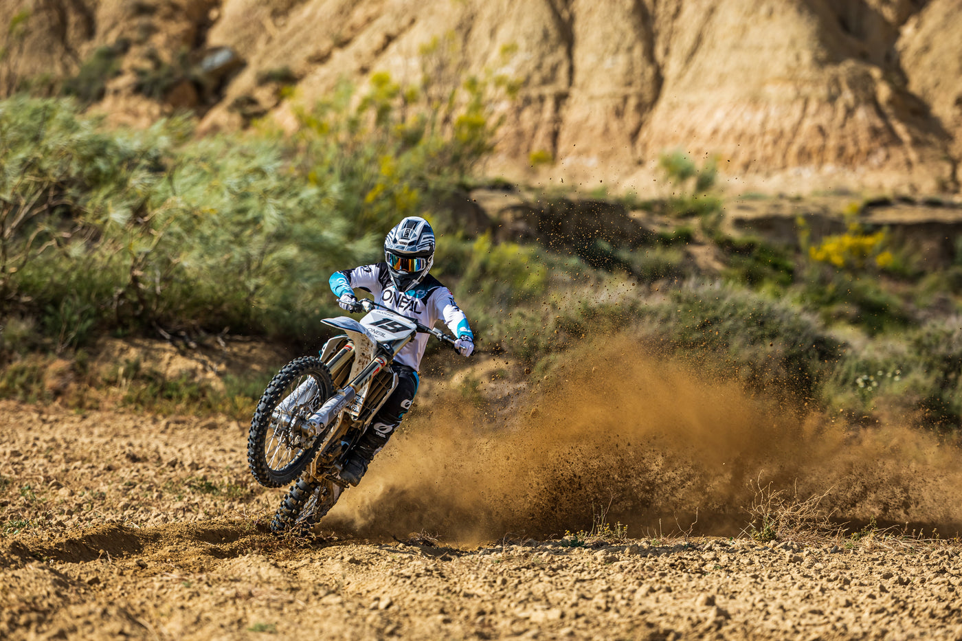 Ein Motocrossfahrer macht auf einem Dirtbike einen Wheelie und wirbelt dabei vor einer schroffen Bergkulisse Staub auf. Der Fahrer trägt die komplette Ausrüstung, einschließlich Helm und Motocross-Bekleidung, auf einer Dirtpiste.