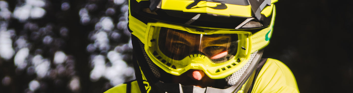 Nahaufnahme einer Person mit einem leuchtend gelben Motocross-Helm und einer Schutzbrille und konzentriertem Gesichtsausdruck. Der dunkle, unscharfe Hintergrund verstärkt den konzentrierten Blick des Fahrers.