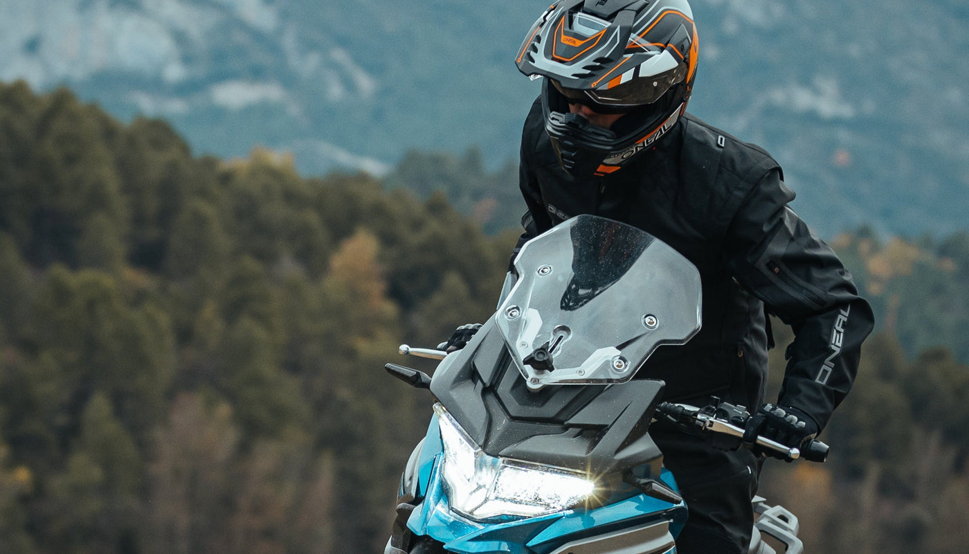 Ein Motorradfahrer mit Helm und schwarzer Ausrüstung fährt eine Straße entlang, im Hintergrund ist ein Bergwald zu sehen.