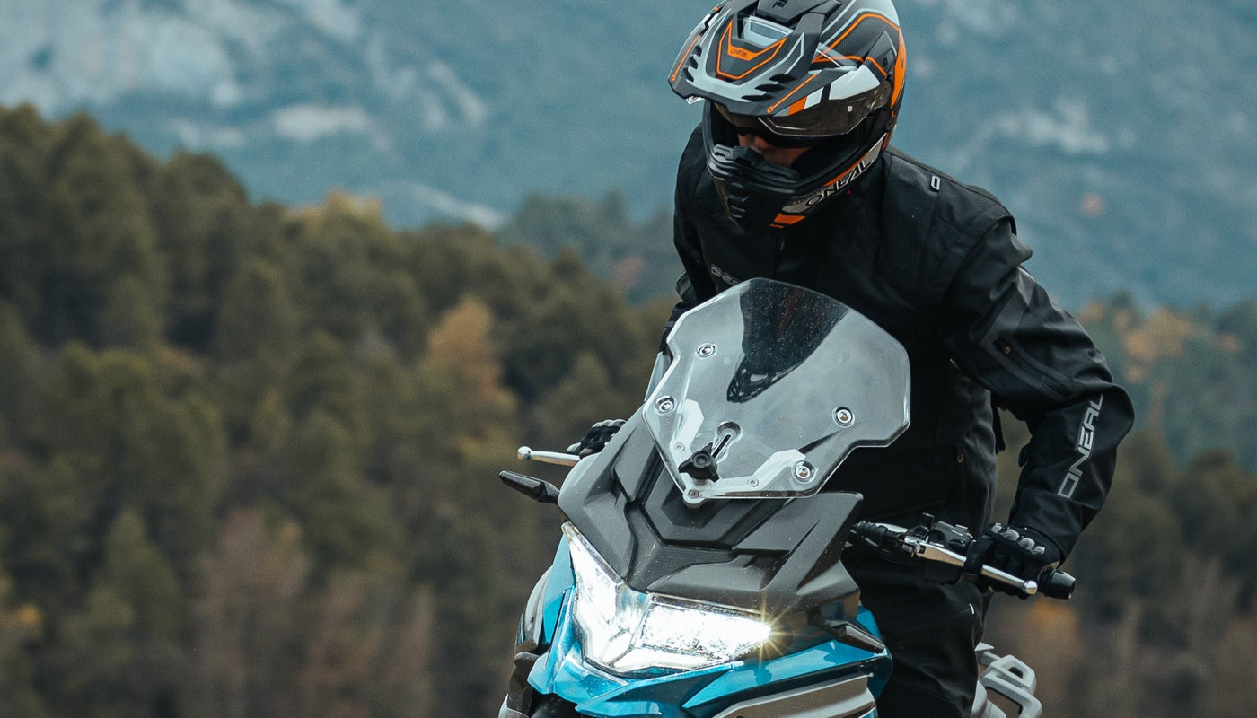 Eine Person mit Helm und schwarzer Jacke fährt mit einem Motorrad durch eine bergige Gegend mit dichten grünen Bäumen im Hintergrund.