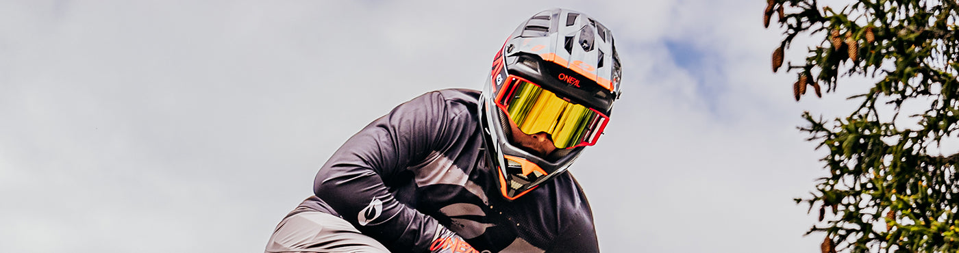 Eine Person mit Integralhelm und Schutzbrille fährt vor einem bewölkten Himmel mit dem Fahrrad bergab. Auf der rechten Seite sind grüne Blätter zu sehen.