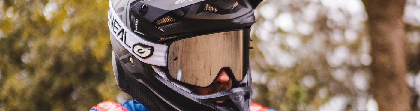 Eine Person mit einem schwarz-weißen Helm und reflektierendem Visier steht vor einem verschwommenen, blätterreichen Hintergrund. Sie trägt ein blau-rotes Outfit und sieht aus, als wäre sie bereit für eine Radtour oder ein Rennen im Freien.