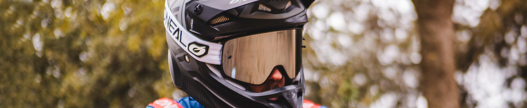 Eine Person mit einem schwarz-weißen Helm und reflektierendem Visier steht vor einem verschwommenen, blätterreichen Hintergrund. Sie trägt ein blau-rotes Outfit und sieht aus, als wäre sie bereit für eine Radtour oder ein Rennen im Freien.