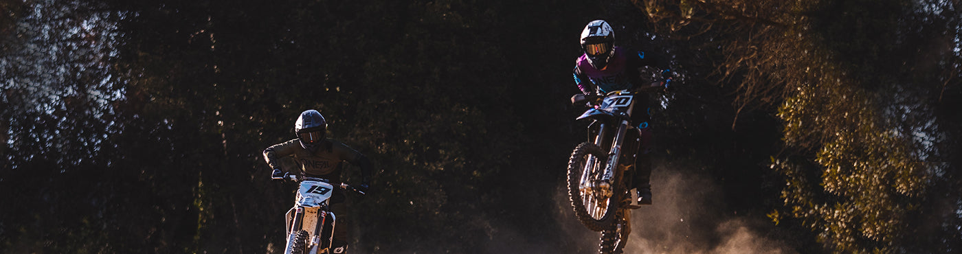 Zwei Motocrossfahrer in der Luft vor einer Baumkulisse. Ein Fahrer links mit weißem Motorrad, Helm und dunkler Ausrüstung; der Fahrer rechts trägt ein buntes Outfit, beide wirbeln Staub auf.