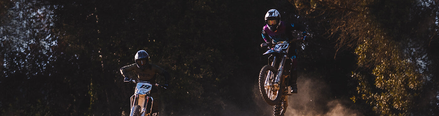 Zwei Motocross-Fahrer in der Luft, einer im dunklen Outfit, der andere in bunter Ausrüstung, fahren ein Rennen auf einer von Bäumen umgebenen Sandpiste, während unter klarem Himmel eine Staubwolke aufsteigt.