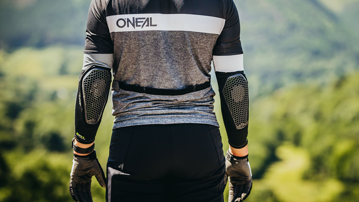 Eine Person in Radsportbekleidung mit Ellbogenschützern und Handschuhen steht vor einer malerischen grünen Landschaft. Auf dem Rücken des Trikots steht das Wort „ONEAL“.