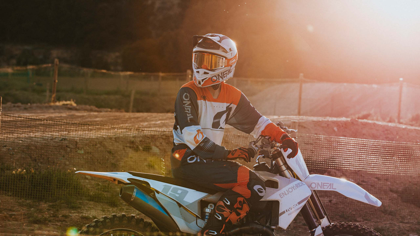 Ein Motocrossfahrer in voller Montur sitzt auf einem Dirtbike und wird dabei von der Sonne auf einer Dirtpiste gebadet. Der Fahrer trägt einen Helm und ein farbenfrohes Outfit, auf dem das Wort „O'NEAL“ zu sehen ist.