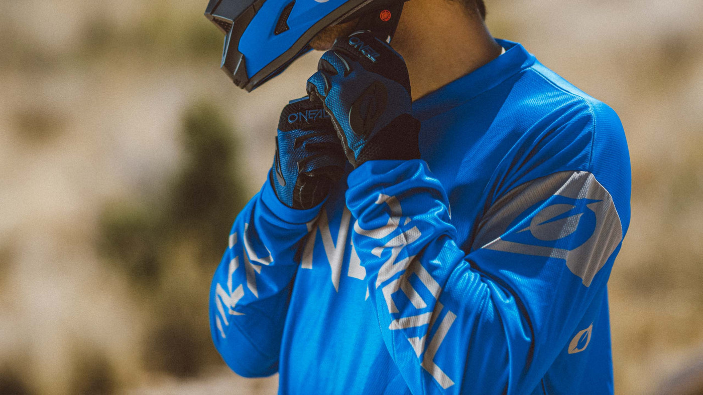 Eine Person mit blauem Motocrosshelm und Handschuhen passt ihren Kinnriemen an. Ihr langärmeliges blaues Trikot weist ein auffälliges weißes Markenlogo auf. Der Hintergrund ist verschwommen und weist einen Hauch von Grün und neutralen Tönen auf.