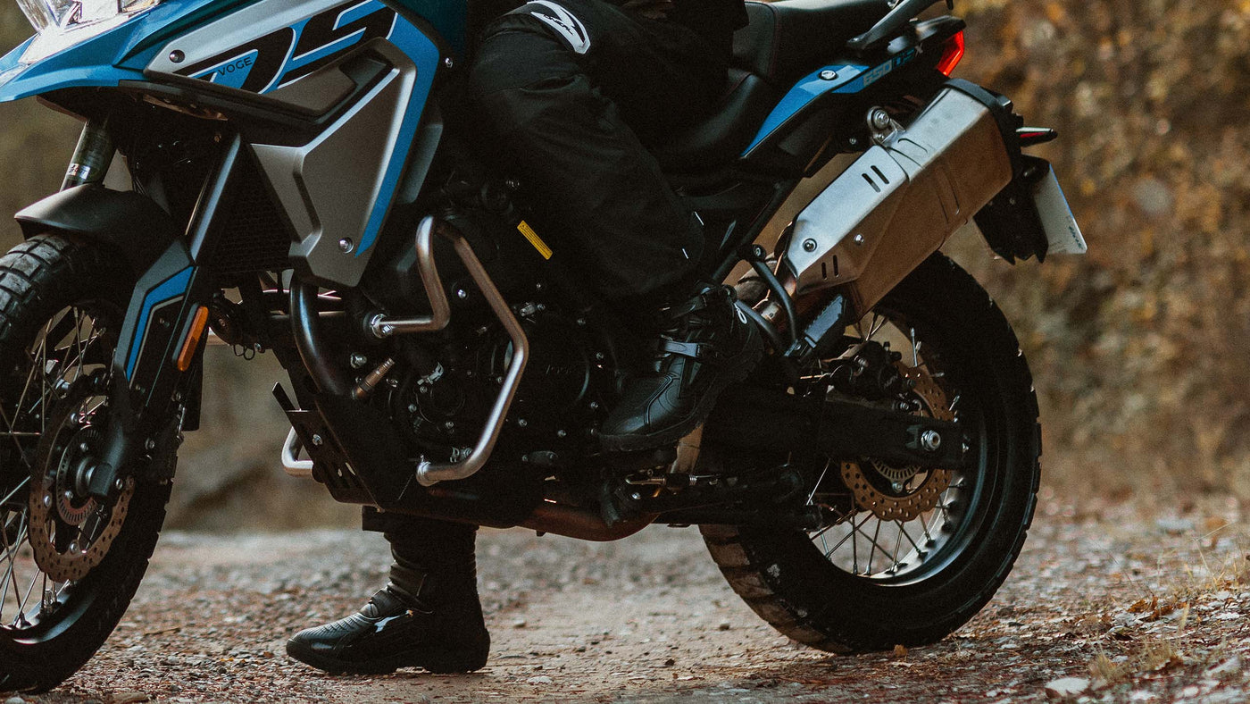 Person auf einem blauen Motorrad mit schwarzer Ausrüstung, geparkt auf einem Schotterweg, umgeben von trockenem Laub.