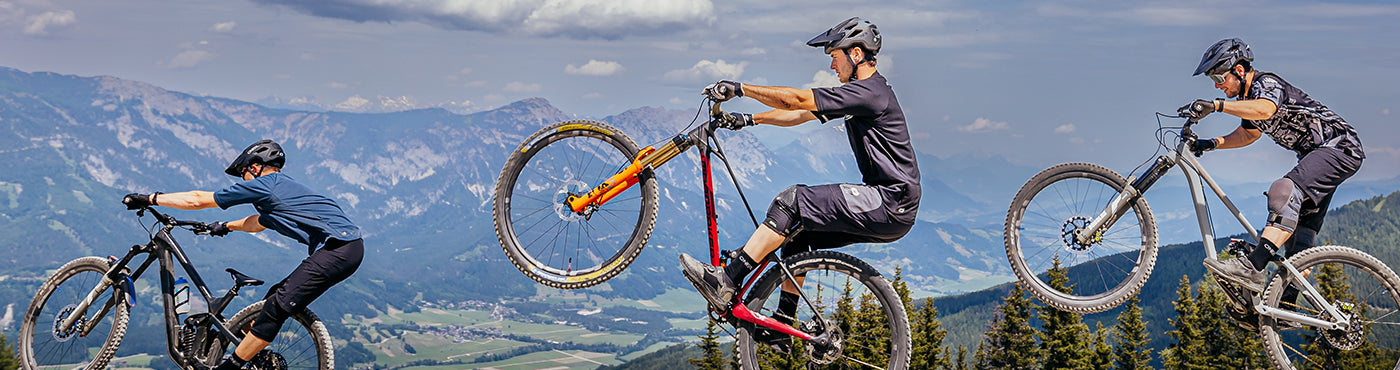 Drei Mountainbiker fahren einen Bergrücken entlang, mit malerischer Aussicht auf Berge und Täler im Hintergrund. Sie fahren in einer Reihe, wobei der mittlere Biker einen Wheelie macht. Der Himmel ist teilweise bewölkt.