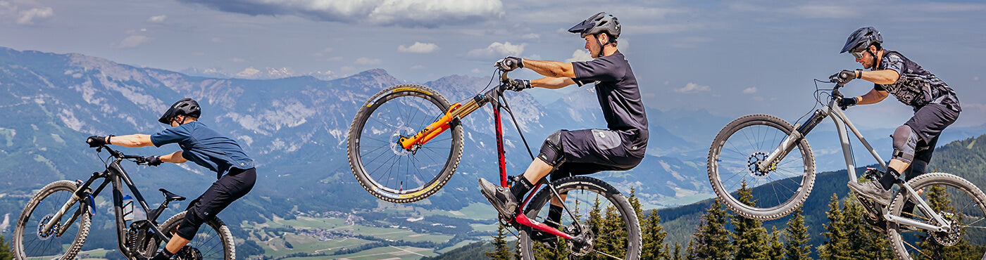 Drei Radfahrer führen vor der Kulisse eines bergigen Geländes und eines teilweise bewölkten Himmels Stunts auf Mountainbikes vor. Sie befinden sich in der Luft und demonstrieren Beweglichkeit und Gleichgewicht inmitten der weitläufigen Naturlandschaft.