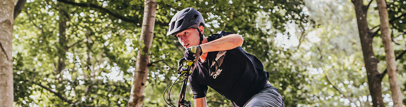 Ein Mountainbiker mit Helm und Handschuhen fährt auf einem Waldweg, umgeben von hohen Bäumen und durch deren Blätter gesprenkeltes Sonnenlicht fällt.