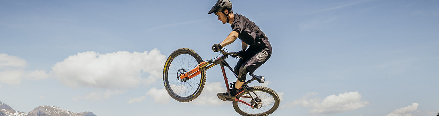 Person, die einen Stunt in der Luft auf einem Mountainbike ausführt und dabei einen Helm und Schutzausrüstung trägt. Im Hintergrund ist ein klarer blauer Himmel mit ein paar Wolken und in der Ferne sind Berge zu sehen.