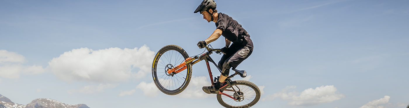 Eine Person mit Helm macht vor einem klaren blauen Himmel einen Sprung auf einem Mountainbike. Im Hintergrund sind schneebedeckte Berge und flauschige Wolken zu sehen.