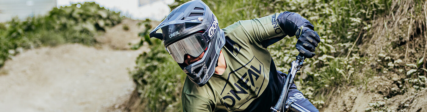 Ein Mountainbiker mit Helm und Schutzbrille fährt einen Feldweg hinunter. Der Weg ist von Grünflächen und Hängen umgeben.