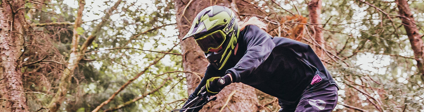 Ein Mountainbiker mit grünem Helm fährt durch einen Waldweg. Die Person ist konzentriert und nach vorne gebeugt und navigiert zwischen hohen Bäumen hindurch. Die Szene ist dynamisch und actionreich.