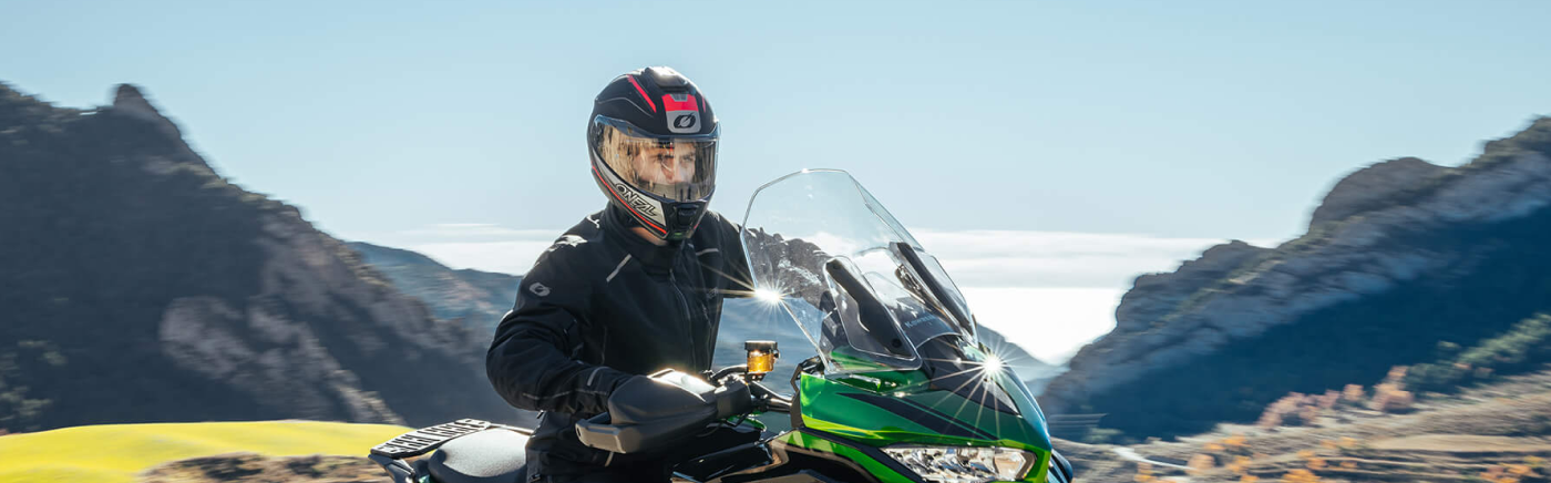 Eine Person mit Helm fährt auf einem grünen Motorrad durch eine Berglandschaft unter einem klaren blauen Himmel. Im Hintergrund sind Felsformationen und ein Feld zu sehen.