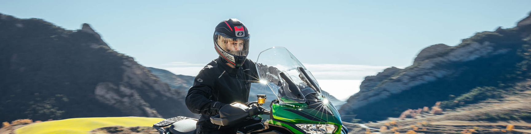 Eine Person mit Helm fährt auf einem grünen Motorrad durch eine Berglandschaft unter einem klaren blauen Himmel. Im Hintergrund sind Felsformationen und ein Feld zu sehen.