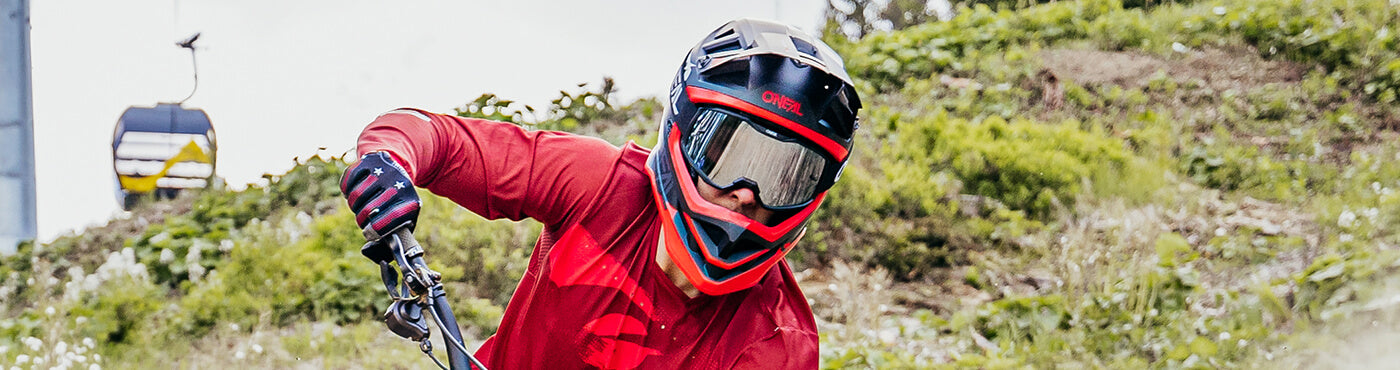 Ein Mountainbiker in einem roten Trikot und einem schwarzen Helm mit Schutzbrille fährt bergab, im Hintergrund sind Skilifte und Grün zu sehen.