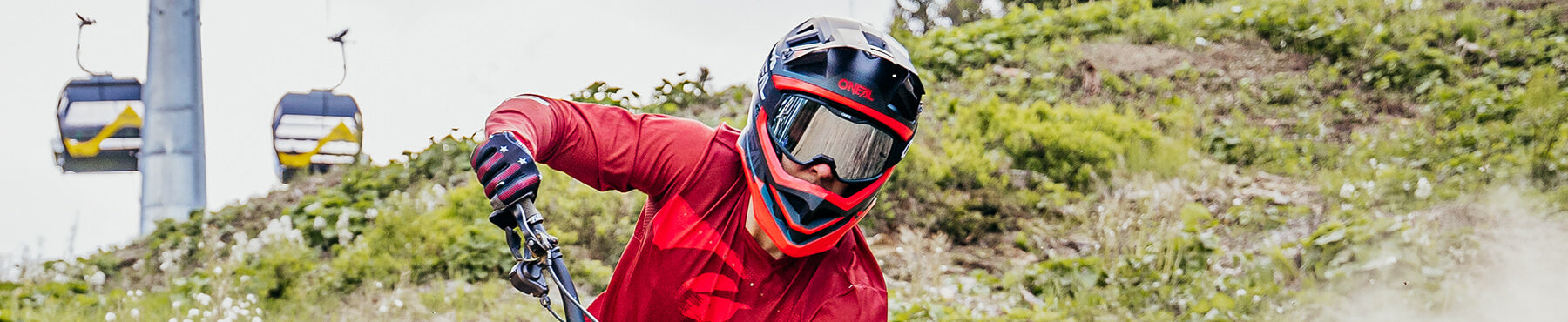 Ein Mountainbiker in einem roten Trikot und einem schwarzen Helm mit Schutzbrille fährt bergab, im Hintergrund sind Skilifte und Grün zu sehen.