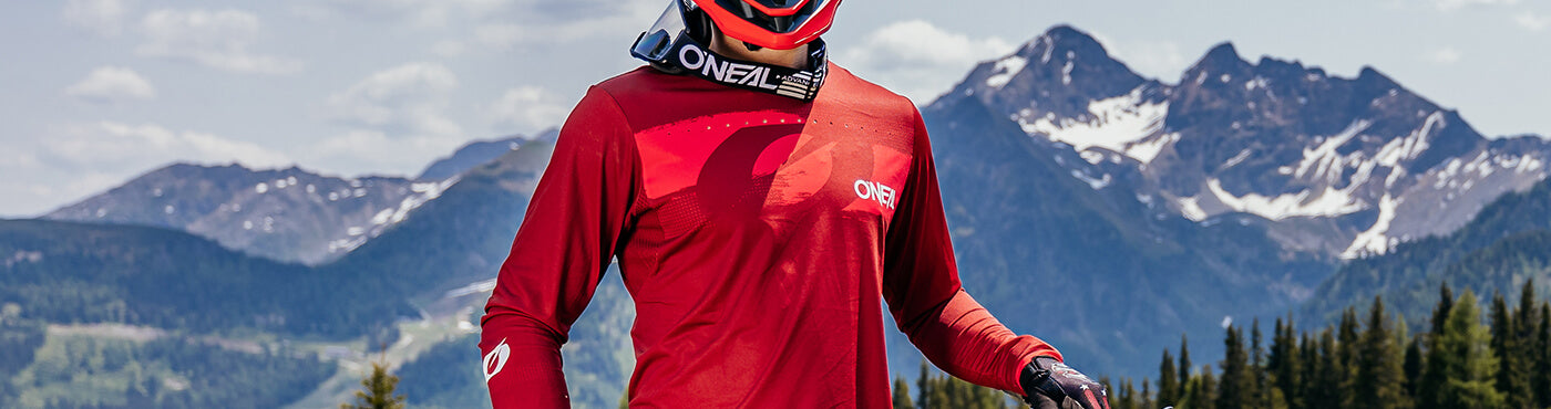 Eine Person in roter Fahrradkleidung und Helm hält ein Mountainbike auf einem Pfad. Im Hintergrund sind schneebedeckte Berge und Kiefern unter einem klaren Himmel zu sehen.