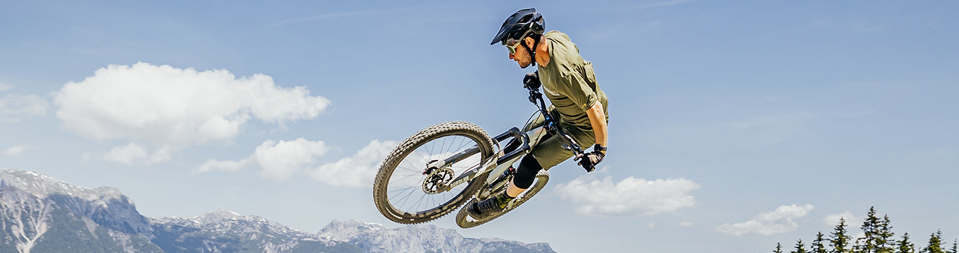 Eine Person mit Helm führt auf einem Mountainbike vor dem Hintergrund eines klaren blauen Himmels und schneebedeckter Berggipfel einen Hochsprung in der Luft aus.