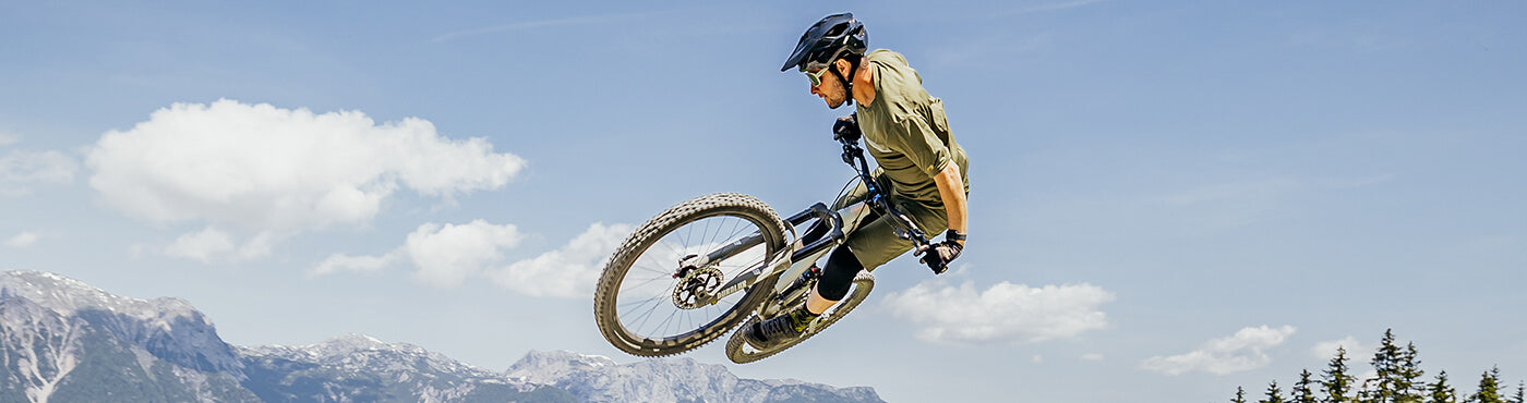 Eine Person mit Helm und Schutzausrüstung führt auf einem Mountainbike vor dem Hintergrund eines klaren blauen Himmels, Wolken und entfernter Berggipfel einen Trick in der Luft aus. Im Vordergrund sind bewaldete Hügel zu sehen.