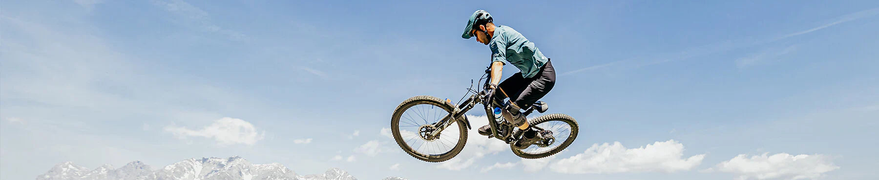 Eine Person mit Helm macht vor einem klaren blauen Himmel einen Sprung in der Luft auf einem Mountainbike. Im Hintergrund sind schneebedeckte Berge zu sehen.