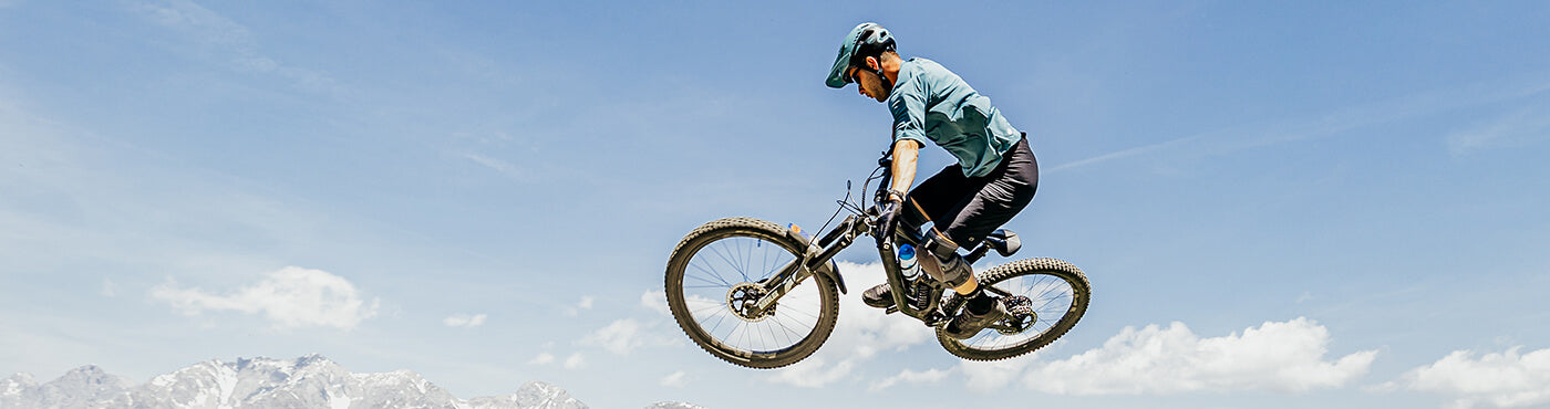 Eine Person in einem blauen Hemd und Helm fährt mit einem Mountainbike durch die Luft vor einem klaren blauen Himmel. In der Ferne sind darunter Berge zu sehen.