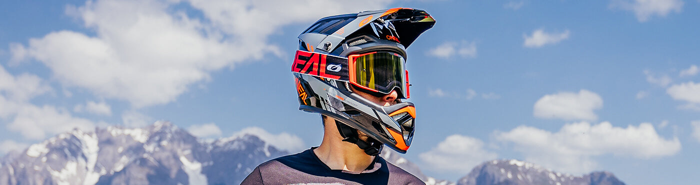 Eine Person mit Motocross-Helm und Schutzbrille steht vor einer Bergkulisse und einem blauen Himmel mit vereinzelten Wolken.