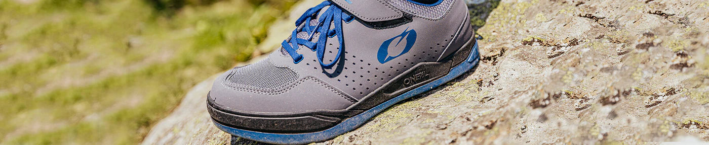 Nahaufnahme eines grau-blauen Sportschuhs mit blauen Schnürsenkeln, der auf einer Felsoberfläche im Freien liegt. Der Schuh verfügt über ein perforiertes Design zur Belüftung und erscheint in einer natürlichen, sonnenbeschienenen Umgebung.