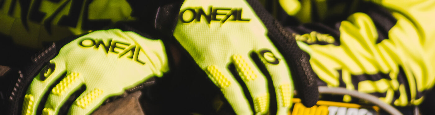 Nahaufnahme einer Person, die leuchtend gelbe Handschuhe mit dem Markennamen „ONEAL“ trägt. Die Handschuhe liegen auf einer Oberfläche, möglicherweise Teil eines Motorrads oder einer Ausrüstung.