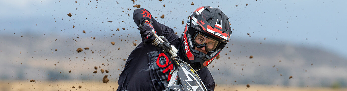 Ein Motocrossfahrer mit Helm und Schutzbrille lehnt sich in eine Kurve und wirbelt dabei Staub über die Strecke. Im Hintergrund ist eine verschwommene Landschaft mit Hügeln unter einem bewölkten Himmel zu sehen.