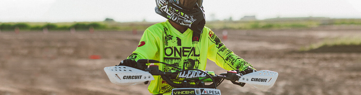 Ein Motocross-Fahrer in neongrüner Ausrüstung und Helm steht auf einem Dirtbike in einem sandigen Gelände, mit verschwommener Naturlandschaft im Hintergrund.