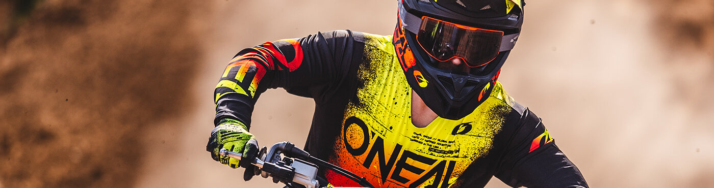 Ein Motocross-Fahrer in farbenfroher Schutzausrüstung und Helm rast umgeben von Staubwolken über eine unbefestigte Strecke.