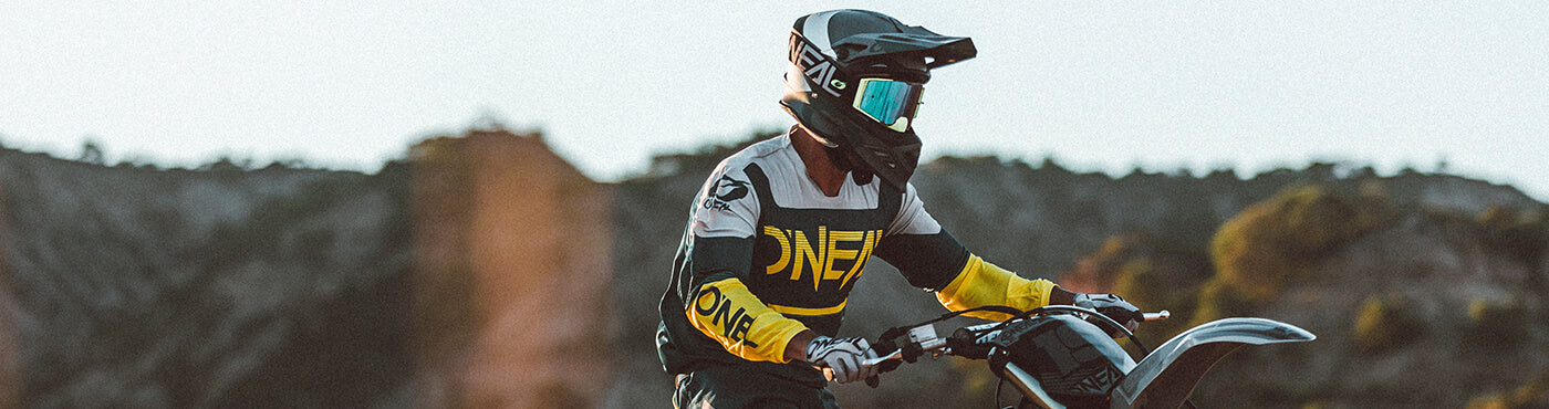 Eine Person in Motocross-Ausrüstung, einschließlich Helm und Schutzbrille, fährt an einem sonnigen Tag ein Dirtbike. Im Hintergrund sind felsige Hügel und ein klarer Himmel zu sehen.