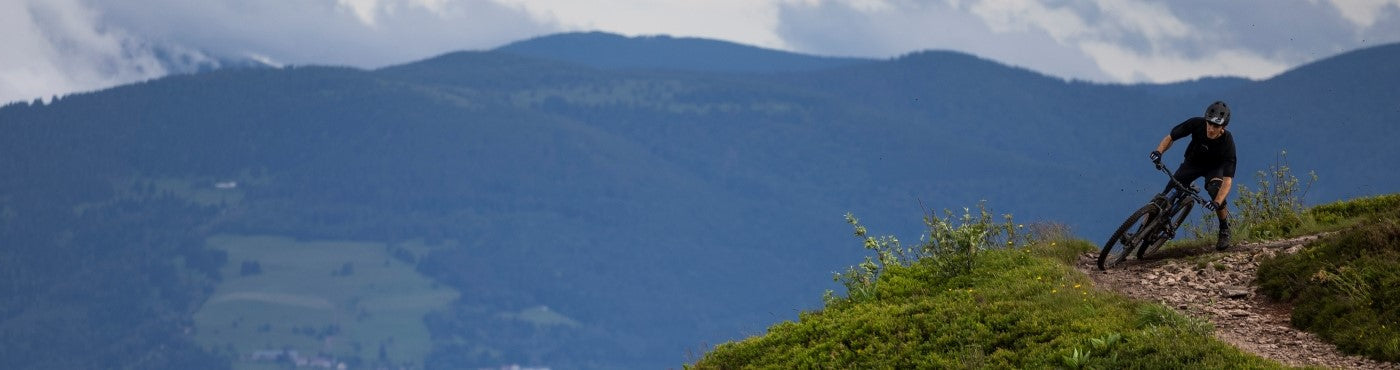 Eine Person fährt mit dem Mountainbike auf einem schmalen Pfad auf einem grünen Hügel, mit Blick auf ausgedehnte bewaldete Berge und einen bewölkten Himmel im Hintergrund.