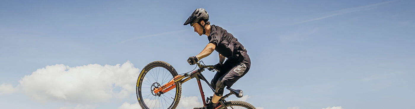 Eine Person macht auf einem Mountainbike einen Wheelie vor einem klaren blauen Himmel mit ein paar Wolken. Sie trägt einen Helm und Radsportbekleidung.