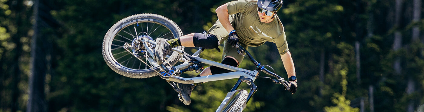 Ein Mountainbiker führt auf einem Pfad mit Waldhintergrund einen Lufttrick aus. Der Fahrer trägt einen Helm, eine Sonnenbrille, Knieschützer und ein grünes Outfit. Das Fahrrad ist in der Luft geneigt.