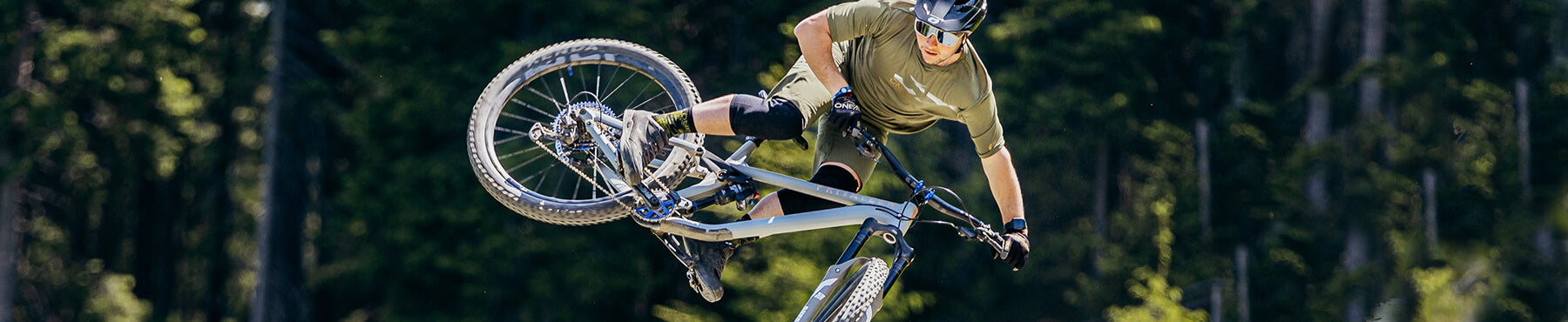 Ein Mountainbiker führt auf einem Pfad mit Waldhintergrund einen Lufttrick aus. Der Fahrer trägt einen Helm, eine Sonnenbrille, Knieschützer und ein grünes Outfit. Das Fahrrad ist in der Luft geneigt.