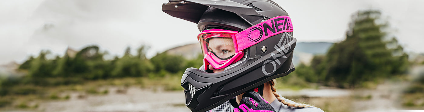 Person mit schwarzem Helm mit rosa Visier und Handschuhen, Blick nach links. Im Hintergrund ist eine natürliche Umgebung im Freien mit verschwommenen Bäumen zu sehen.