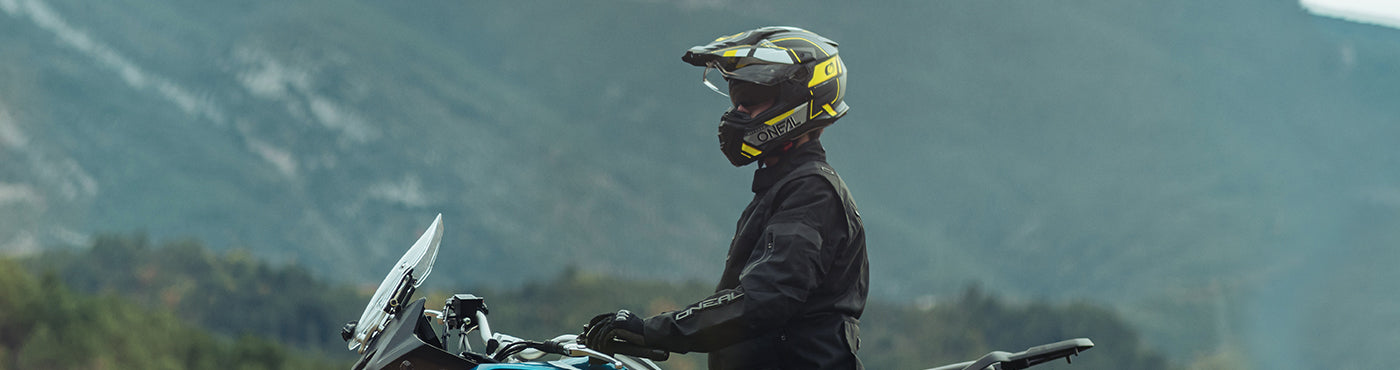 Motorradfahrer in schwarzer Montur und Helm sitzt auf einem Motorrad mit Bergen im Hintergrund. Der Fahrer blickt nach links und ist bereit, durch die malerische Landschaft zu fahren.