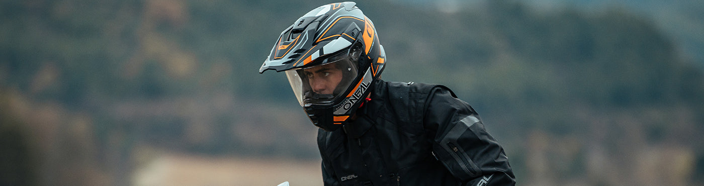 Eine Person mit einem schwarz-orangen Motorradhelm und einer schwarzen Jacke steht im Freien mit verschwommener Natur im Hintergrund.