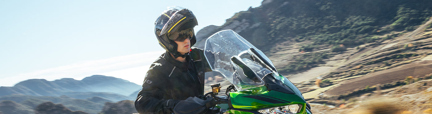 Eine Person mit Helm und schwarzer Jacke fährt auf einem grünen Motorrad durch eine Berglandschaft mit klarem Himmel und zerklüftetem Gelände im Hintergrund.