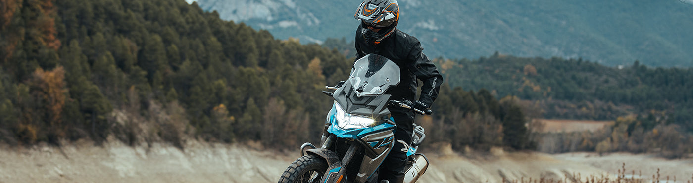 Eine Person in Motorradmontur fährt auf einem Motorrad einen schroffen Pfad entlang, im Hintergrund sind Berge und dichter Wald zu sehen. Der Himmel ist bedeckt, was die abenteuerliche Atmosphäre noch verstärkt.