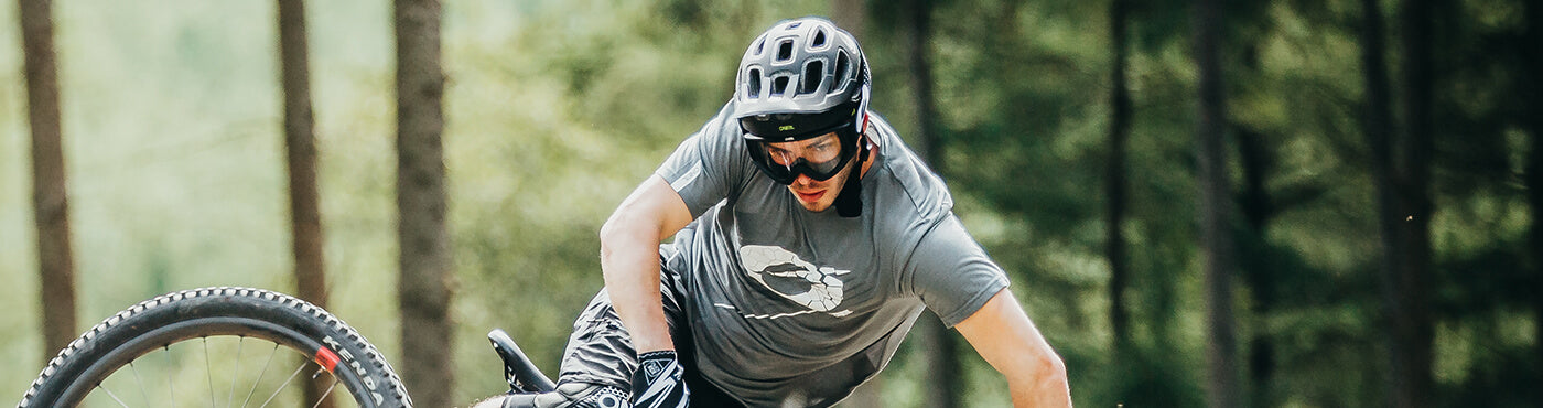 Eine Person mit Helm und Schutzbrille fährt mit einem Mountainbike durch einen Waldweg. Sie trägt ein graues Hemd und schwarze Shorts. Im Hintergrund sind Bäume zu sehen.