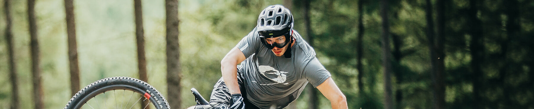 Eine Person mit Helm und Schutzbrille fährt mit einem Mountainbike durch einen Waldweg. Sie trägt ein graues Hemd und schwarze Shorts. Im Hintergrund sind Bäume zu sehen.