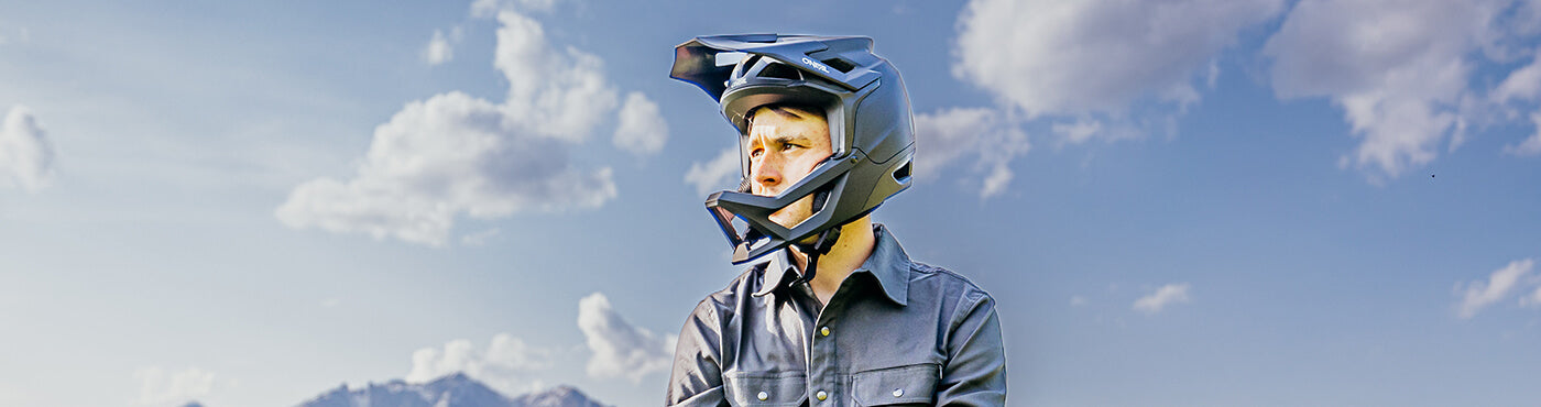 Eine Person mit Integralhelm blickt zur Seite unter einem blauen Himmel mit einigen Wolken. Im Hintergrund sind schwach Berge zu erkennen.